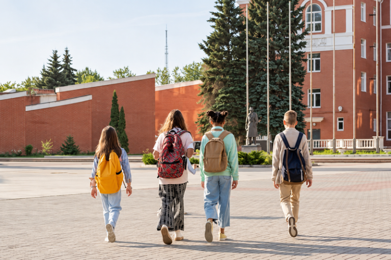 Four young people wearing backpacks walk across a paved courtyard toward a school building with reddish-orange walls and cream trim.