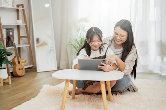 An Asian mother and young daughter sit together on the floor at home, smiling as they use a tablet at a small coffee table.